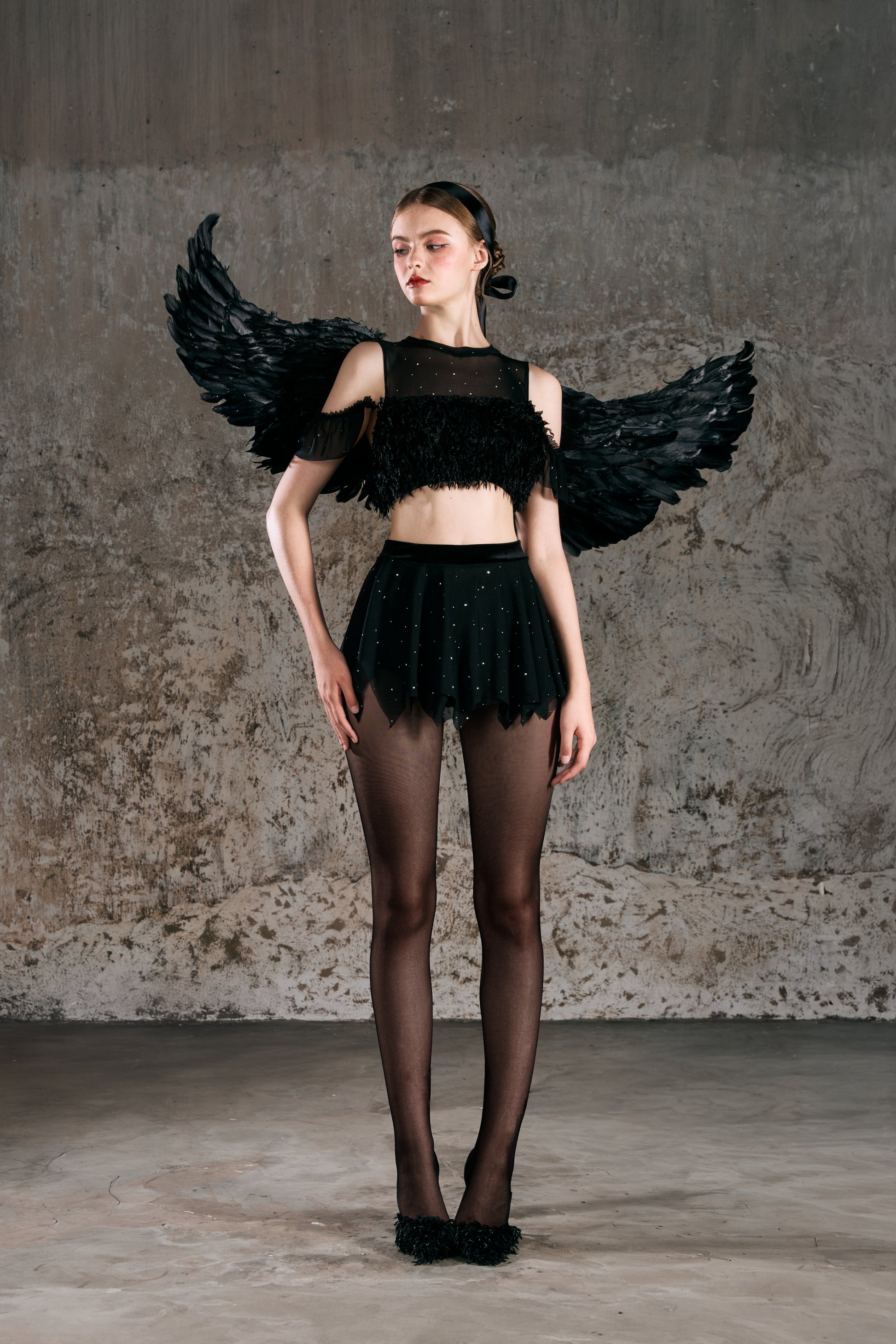Model wearing a black outfit with large wings against a textured wall Hebe designed by Spincess