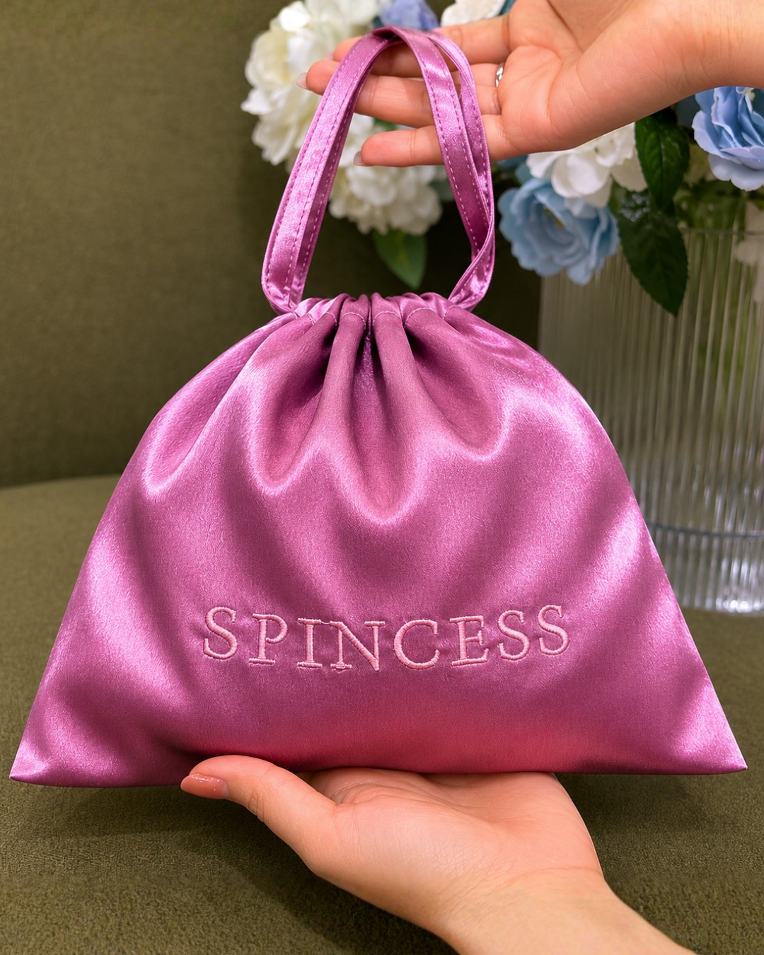 Pink satin drawstring bag with 'SPINCESS' text held by a hand, against a floral background. Satin Bag Pink Designed by Spincess